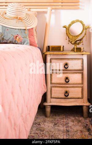 Stylischer Damen-Sonnenhut auf dem Bett neben dem Nachttisch mit Spiegel. Eleganz in femininem Stil. Interieur, Retro Stockfoto