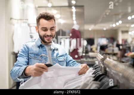 Glücklicher junger Mann mit Hemd in den Händen, der neue Kleidung wählt Im Geschäft Stockfoto
