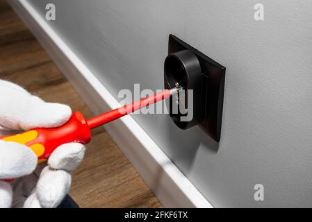 Ein Elektriker verwendet einen roten Schraubendreher, um eine schwarze, doppelt geerdete Steckdose in den Elektrokasten in der Raumwand zu Schrauben. Stockfoto