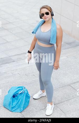 Sportlerin in Übergröße, die im Freien steht, in Sonnenbrillen und Kopfhörern steht und die Kamera anschaut Stockfoto