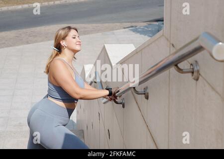 Junge hübsche Plus-Size-Frau, die sich am Treppengeländer im Freien dehnt Und genießen Sie warme Strahlen der Morgensonne Stockfoto