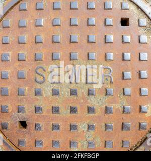 Eine Schachtabdeckung aus schwerem Metall mit dem Wort KANALISATION darauf. Erhabene Buchstaben und quadratische Beulen. Leichter Oberflächenrost. Nahaufnahme von direkt oben. Stockfoto