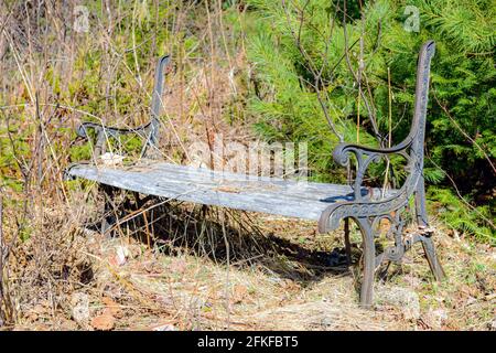 Eine alte, kaputte Parkbank im Wald. Rücken fehlt, Holz verblasst, Metall zeigt Rostflecken. Gras und Bäume um die Bank herum. Sonniger Tag, konzentrieren Sie sich auf den nächsten Stockfoto