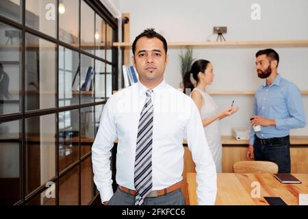 Porträt eines ernsthaften indischen Unternehmers in weißem Hemd, der die Kamera anschaut, während sein Kollege im Hintergrund spricht Stockfoto