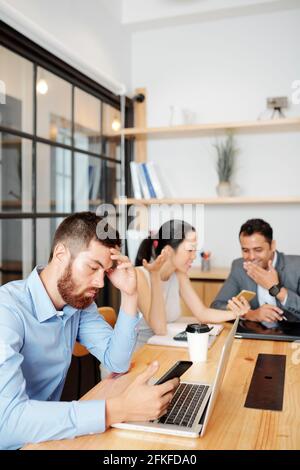 Ernster Mann liest Nachrichten auf dem Smartphone, wenn seine Kollegen diskutieren Frische Meme im Hintergrund während der Kaffeepause Stockfoto