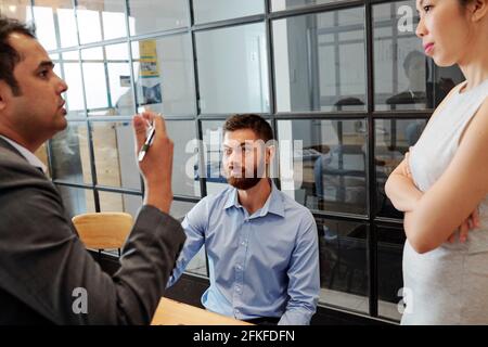 Lächelnder junger bärtiger Geschäftsmann, der seiner emotionalen Rede zuhört Kollege bei der täglichen Besprechung Stockfoto