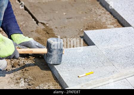 Ein Gummihammer in den Händen eines Maurers vor dem Hintergrund der Verlegung von Granit Pflasterplatten an einem sonnigen Tag. Stockfoto