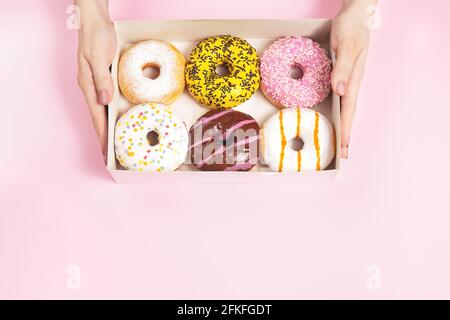 Weibliche Hände halten eine Schachtel mit glasierten Donuts auf einem pastellrosa Hintergrund. Konzept Süßwarenladen, Gebäck, Café. Draufsicht, flach liegend, Kopie sp Stockfoto