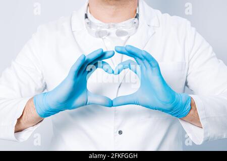 Doktor Hände in Handschuhen in der Herzform vor dem Hintergrund seines Körpers und medizinischen Kleides. Nahaufnahme. Männliche Hand in blauen medizinischen Handschuhen zeigt Herz Stockfoto