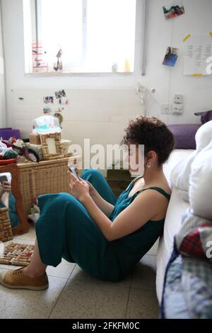 Die schöne 21-jährige Frau mit wunderschönen lockigen Haaren liest Textnachrichten auf ihrem Handy, während sie auf dem Boden in ihrem Zimmer gegen ihr Bett sitzt. Stockfoto