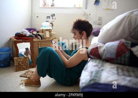 Schöne 21-jährige Frau mit wunderschönen lockigen Haaren lächelt Und liest gerne Textnachrichten auf ihrem Handy, während sie Sitzt auf dem Boden in ihrem Zimmer Stockfoto