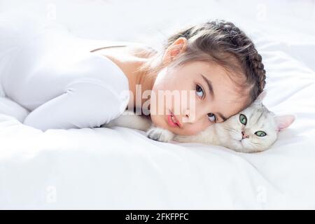 Porträt eines schönen kleinen Mädchens in weißen Kleidern, liegt auf einem sauberen, weißen Bett mit einer weißen britischen Katze. Stockfoto