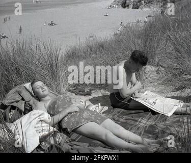 1960er Jahre, historisch, ein Paar, das sich auf einem Teppich auf einigen Sanddünen an einem Sandstrand entspannt, die Dame, die sich sonnt, der Mann, der eine Zeitung liest, England, Großbritannien. Stockfoto