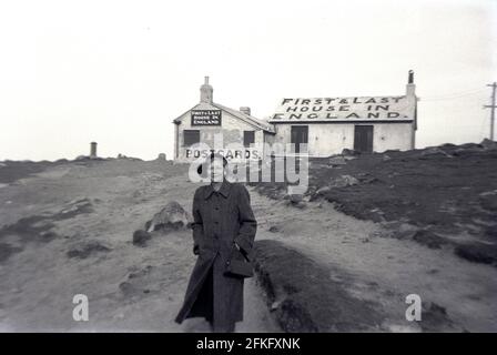 1950er Jahre, historisch, Lands End, Cornwall und eine Dame, die vor einem alten Cottage, dem First & Last House in England, für ein Foto steht - wie in großen Schriftzügen auf dem Dach geschrieben ist - eine berühmte Teestube und ein Souvenirladen an der zerklüfteten Westküste Englands. Da die alte einstöckige Hütte heruntergekommen aussieht und die Fenster zu dieser Zeit vertafelt sind, war sie für den Winter geschlossen. Stockfoto