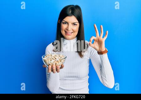 Junge hispanische Frau hält Kürbiskerne Schüssel tun ok Zeichen mit den Fingern, lächelnd freundlich gestikulieren ausgezeichnetes Symbol Stockfoto