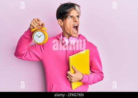 Junger hispanischer Mann hält Buch und Wecker wütend und wütend schreiend frustriert und wütend, schreiend vor Wut. Wut und aggressives Konzept. Stockfoto