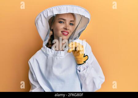 Schöne blonde kaukasische Frau trägt schützende Imkeruniform sieht zuversichtlich aus An der Kamera lächelnd mit gekreuzten Armen und Händen Erhöht am Stockfoto