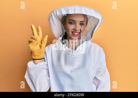 Schöne blonde kaukasische Frau trägt schützende Imker Uniform zeigen und Zeigt mit den Fingern Nummer zwei nach oben und lächelt zuversichtlich und happ Stockfoto