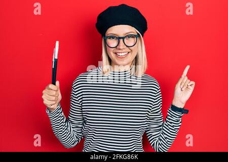 Schöne blonde Frau im französischen Look mit Baskenmütze hält Maler Pinsel lächeln glücklich zeigen mit Hand und Finger auf die Seite Stockfoto