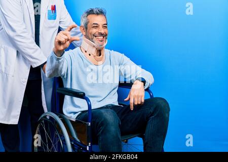 Ein hübscher Mann mittleren Alters mit grauen Haaren auf dem Rollstuhl Halsband lächelnd und selbstbewusst gestikulieren mit kleiner Hand Größenschild mit fing Stockfoto