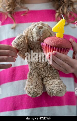 Zum vierten Geburtstag eines Mädchens trägt ein Mädchen in einem rosa-weißen Hemd einen kleinen Teddybären und einen rosa Cupcake mit einer Kerze mit der Nummer Fou Stockfoto