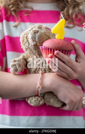 Zum vierten Geburtstag eines Mädchens trägt ein Mädchen in einem rosa-weißen Hemd einen kleinen Teddybären und einen rosa Cupcake mit einer Kerze mit der Nummer Fou Stockfoto