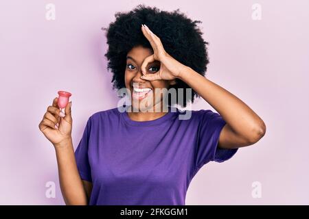 Junge afroamerikanische Frau hält Menstruationstasse lächelnd glücklich tun ok Schild mit Hand auf Auge durch die Finger schauen Stockfoto