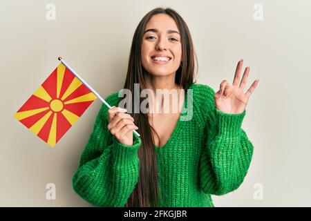 Junge hispanische Mädchen hält mazedonische Flagge tut ok Zeichen mit Fingern, lächelnd freundlich gestikulieren ausgezeichnetes Symbol Stockfoto