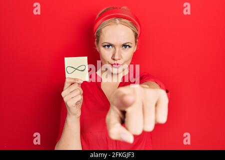 Junge blonde Frau hält unendliche Symbol Erinnerung zeigt mit dem Finger auf die Kamera und Sie, selbstbewusste Geste, die ernst Stockfoto