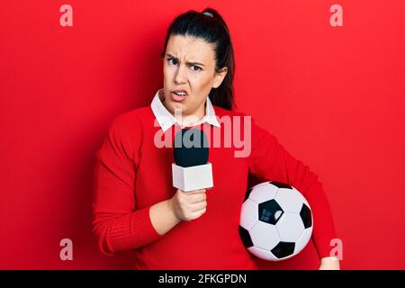 Junge hispanische Frau hält Reportermikrofon und Fußball ahnungslos und verwirrt Ausdruck. Zweifel Konzept. Stockfoto