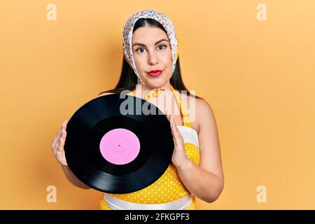 Junge hispanische Frau trägt Pin-up-Stil hält Vinyl-Disc entspannt mit ernstem Ausdruck auf dem Gesicht. Einfach und natürlich Blick auf die Kamera. Stockfoto