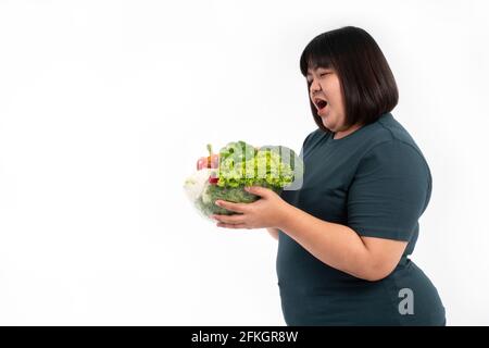 Glückliche übergewichtige asiatische Frau, die einen Gemüskorb hält und auf einem isolierten weißen Hintergrund lächelt, kommt das Konzept der guten Gesundheit vom Essen einer Nutritiou Stockfoto