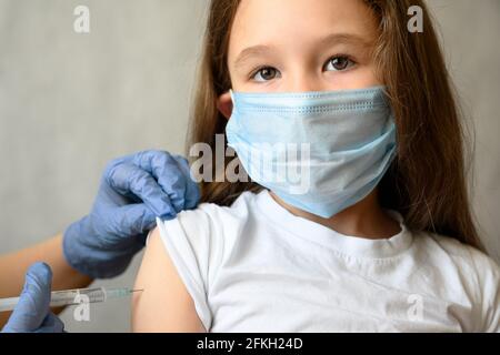 Impfung von Kindern durch COVID-19 oder Grippe, hübsches kleines Mädchen schaut während der Injektion des Coronavirus-Impfstoffs auf die Kamera. Niedliches Kindergesicht in Maske und Spritze. Stockfoto