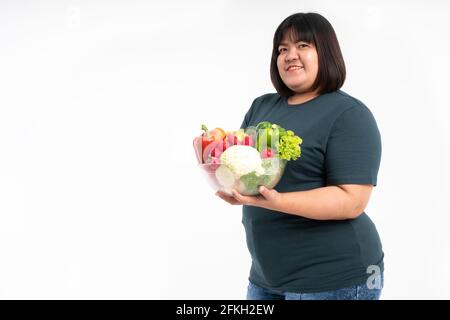 Glückliche übergewichtige asiatische Frau, die einen Gemüskorb hält und auf einem isolierten weißen Hintergrund lächelt, kommt das Konzept der guten Gesundheit vom Essen einer Nutritiou Stockfoto