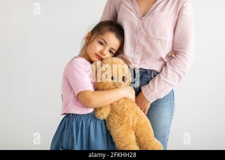 Nettes kleines Mädchen mit Teddybär umarmt Mutter und stehend Zusammen gegen eine leichte Wand Stockfoto