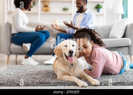 Traurige schwarze Mädchen umarmen Hund, Eltern kämpfen im Hintergrund Stockfoto