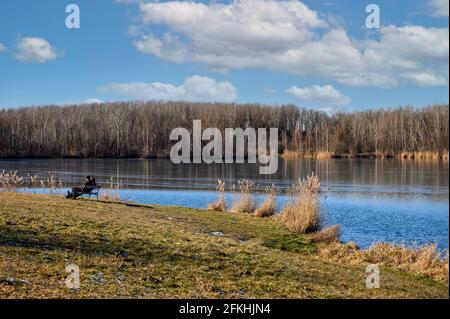 Ein Paar sitzt auf einer Bank und genießt die ersten Strahlen der Frühlingssonne auf einem Flussufer. Stockfoto