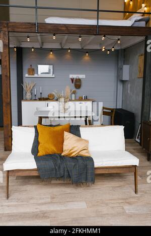 Stilvolles und skandinavisches Interieur des Wohnzimmers einer modernen Wohnung mit einem weißen Sofa, Kissen, einer Decke, einer Küche im Hintergrund. Modern Stockfoto