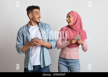 Online-Angebot. Aufgeregt islamisches Paar hält Smartphones und schaut sich gegenseitig an Stockfoto