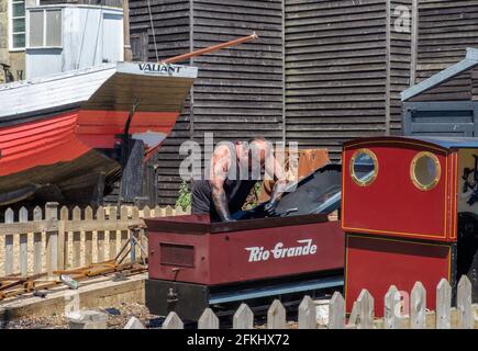 Ein Mann mit schwarzem Fett an den Armen arbeitet am Motor der Miniatureisenbahn am Stade, Hastings, Old Valiant Fischerboot & Net Shops im Hintergrund. Stockfoto