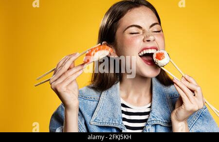 Glückliches Mädchen isst Sushi mit Zufriedenheit, hält Sashimi und beißt Sushi-Rolle mit Lächeln, bestellen köstliche Takeaway in Smartphone-App, zufrieden mit schnell Stockfoto
