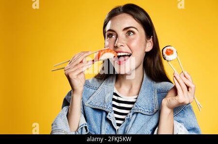 Glückliche Frauen essen Sashimi mit Essstäbchen. Mädchen bestellt Fast-Food-Lieferung aus japanischem Restaurant, hält makizushi Lieblings-Sushi-Rollen und lächelt Stockfoto