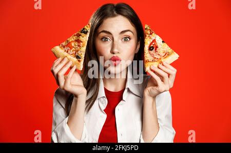 Nettes Mädchen zeigt zwei Scheiben von leckeren Pizza zum Mitnehmen, so dass küssende Gesicht, liebt Pizzeria Fastfood, genießt die Bestellung Lieferung von Lebensmitteln zu Hause, Essen Stockfoto