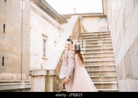 Braut umarmt lächelnden Bräutigam von hinten. Brautpaar stehen auf den Stufen eines alten Gebäudes in Bergamo, Italien Stockfoto