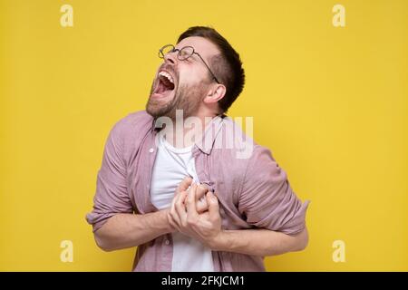 Der junge, attraktive Mann hat starke Schmerzen im Herzen, er hält die Hände auf der Brust und schreit mit geschlossenen Augen. Gelber Hintergrund. Stockfoto