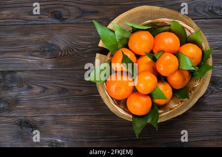 Korb mit frischen Mandarinenorangen, ganz, halbiert und geschält, mit Blättern, auf einem dunkelbraunen Tisch mit Holzstruktur. Gesunde vegane vegetarische Zitrusfrüchte, vi Stockfoto