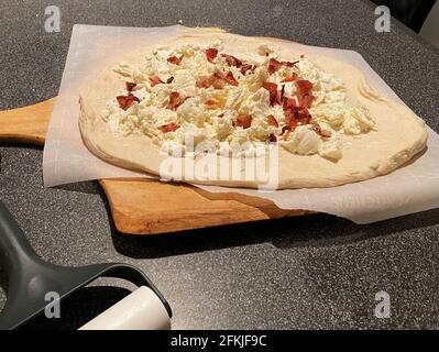 Eine frisch zubereitete, hausgemachte Pizza mit Speck auf Wachspapier auf einem Pizzabrett, das bereit ist, in den Ofen gestellt zu werden. Stockfoto