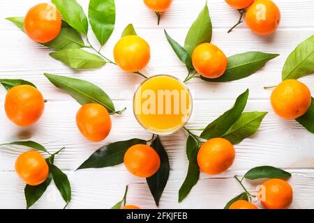 Stilvolle Komposition mit einem Glas Orangensaft & einem Bund frischer Mandarinen mit Blättern auf weißem, strukturiertem Holzhintergrund. Roher Vegetarier Stockfoto