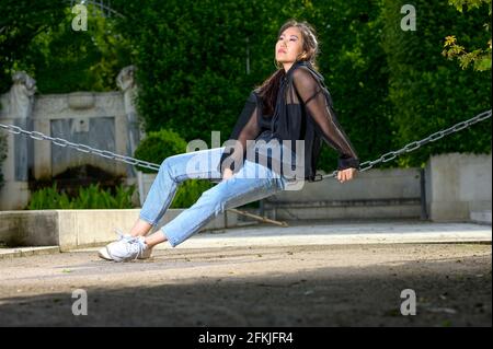 Die hübsche junge mongolische Frau sitzt auf einer dicken Kordel im Park und blickt in die Ferne. Stockfoto