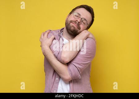 Netter, lustiger bärtiger Mann in runden Gläsern umarmt sich, er träumt von Liebe oder leidet unter Narzissmus, schließt die Augen und lächelt süß. Stockfoto